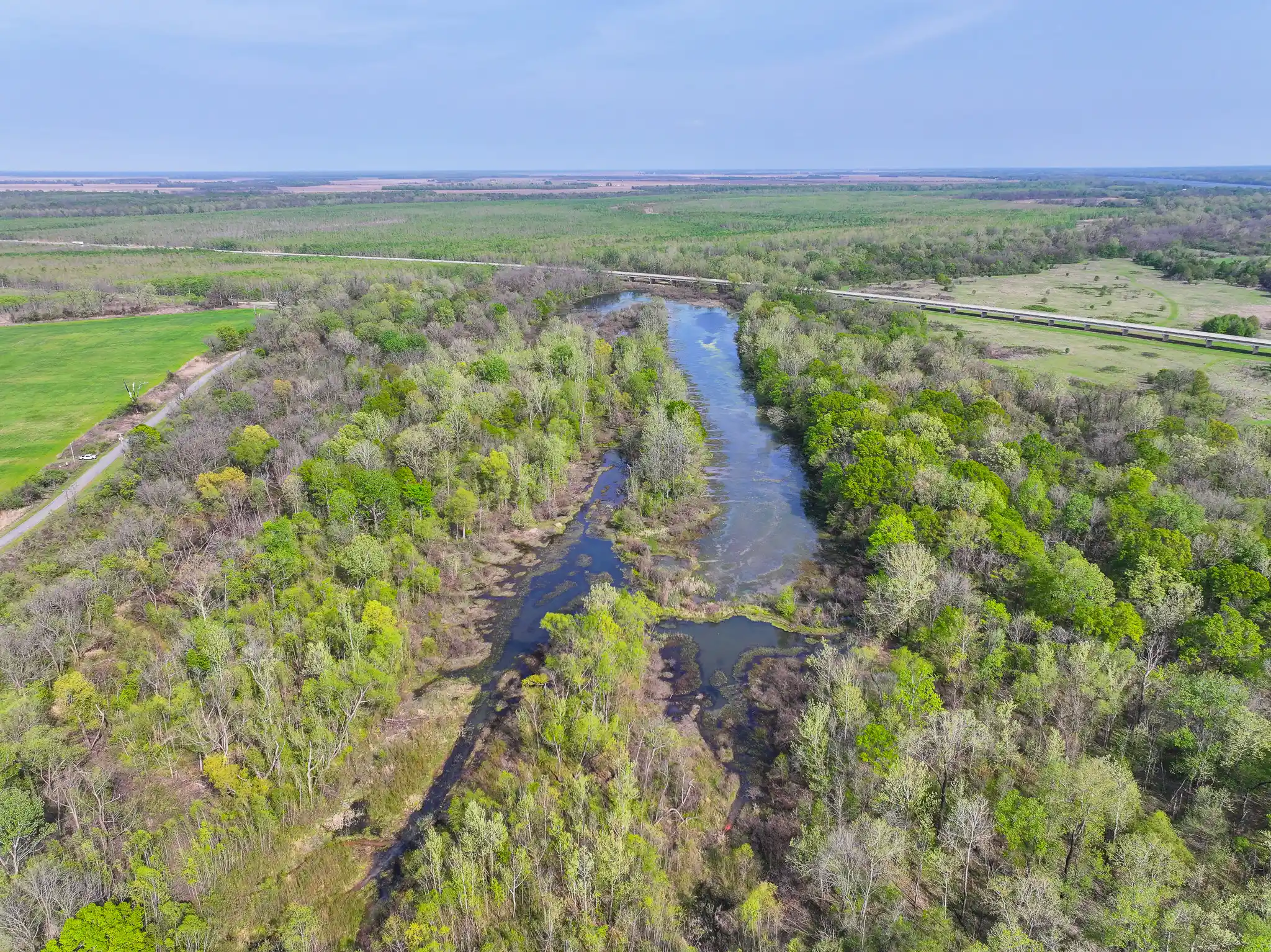 Aerial view of a river winding through a dense, green forest with patches of open land and a road running parallel to the river in the background under a blue sky.