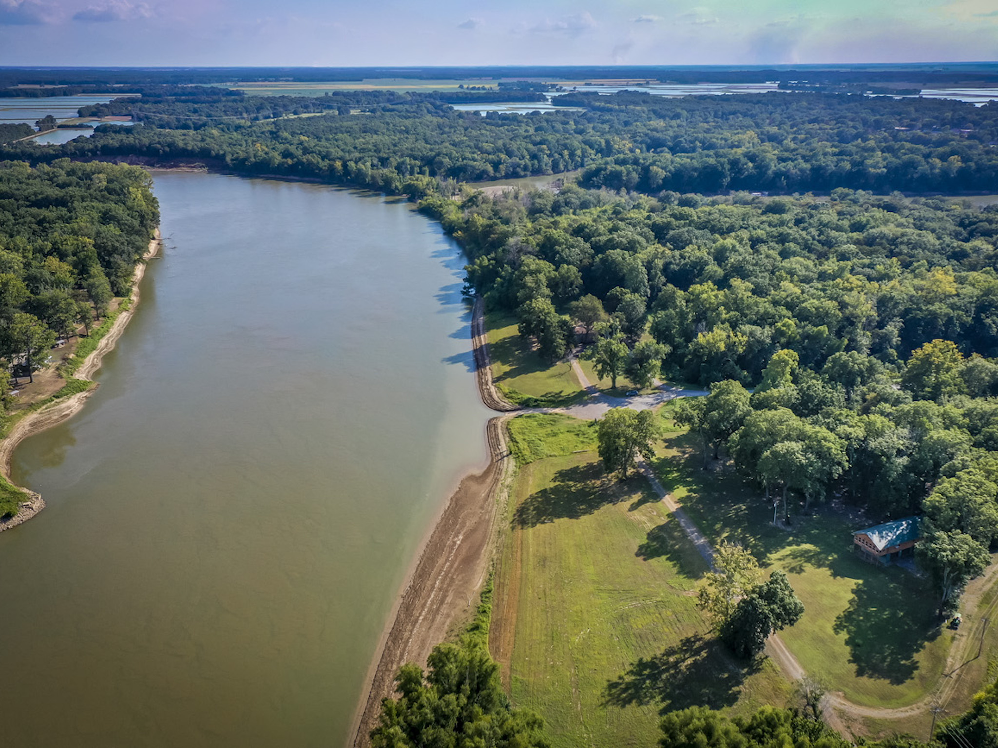 Aerial view of a wide, winding river bordered by dense green forest and grassy open land with a small house; blue sky and distant lakes are visible on the horizon.