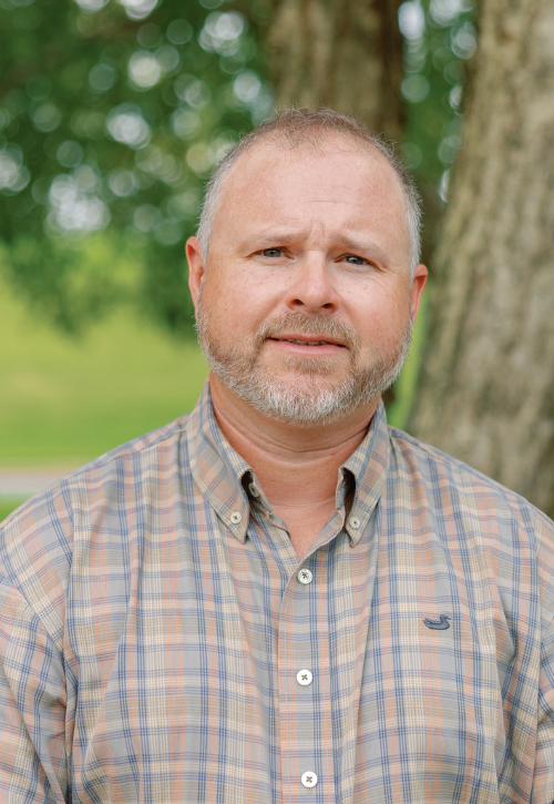 A middle-aged man with short hair and a beard, wearing a plaid button-up shirt, stands outdoors in front of a tree with green foliage in the background.