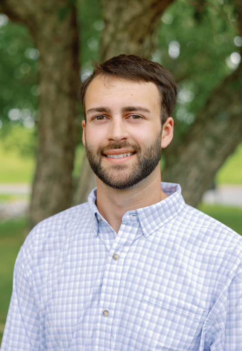 A man with short brown hair and a trimmed beard, wearing a light blue checkered shirt, stands outdoors in front of a tree with green leaves, smiling slightly at the camera.