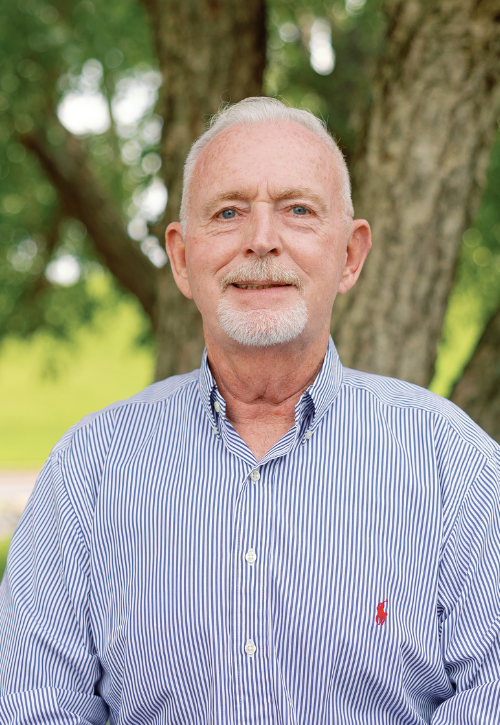 An older man with short white hair and a trimmed white beard is wearing a blue and white striped button-up shirt, standing outdoors in front of a tree with green foliage in the background.