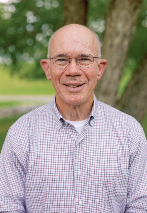 An older man with glasses and a checked shirt smiles at the camera while standing outdoors in front of a tree and blurred greenery.