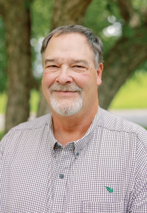A middle-aged man with gray hair and a beard smiles at the camera, wearing a checkered shirt, standing outdoors in front of leafy trees.