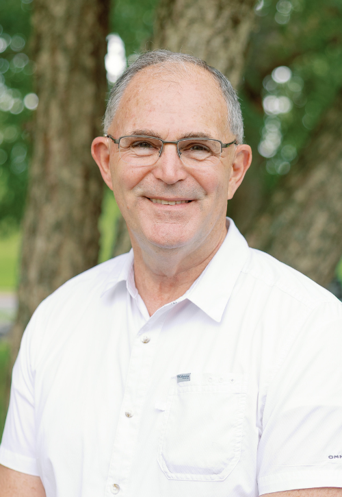 An older man with short gray hair and glasses smiles at the camera. He is wearing a white collared shirt and standing outdoors in front of trees with green leaves.