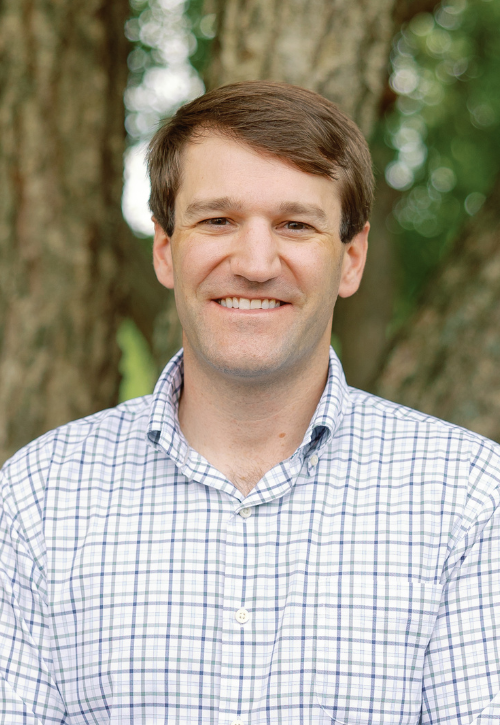 A man with short brown hair, wearing a white and blue checkered shirt, smiles while standing outdoors in front of a tree.