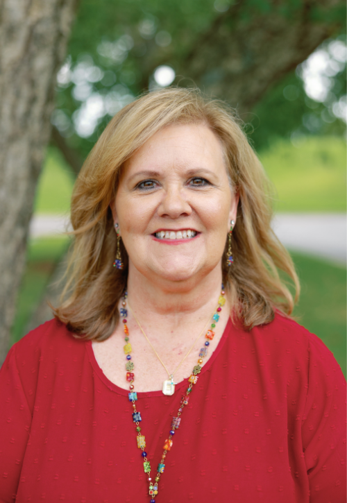 A woman with shoulder-length blonde hair, wearing a red top and colorful beaded necklace, smiles outdoors in front of green trees and a blurred grassy background.