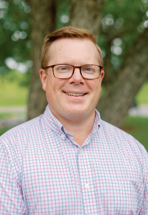 A smiling man with short light brown hair and glasses wears a light pink and blue plaid shirt, standing outdoors in front of a tree with green foliage in the background.