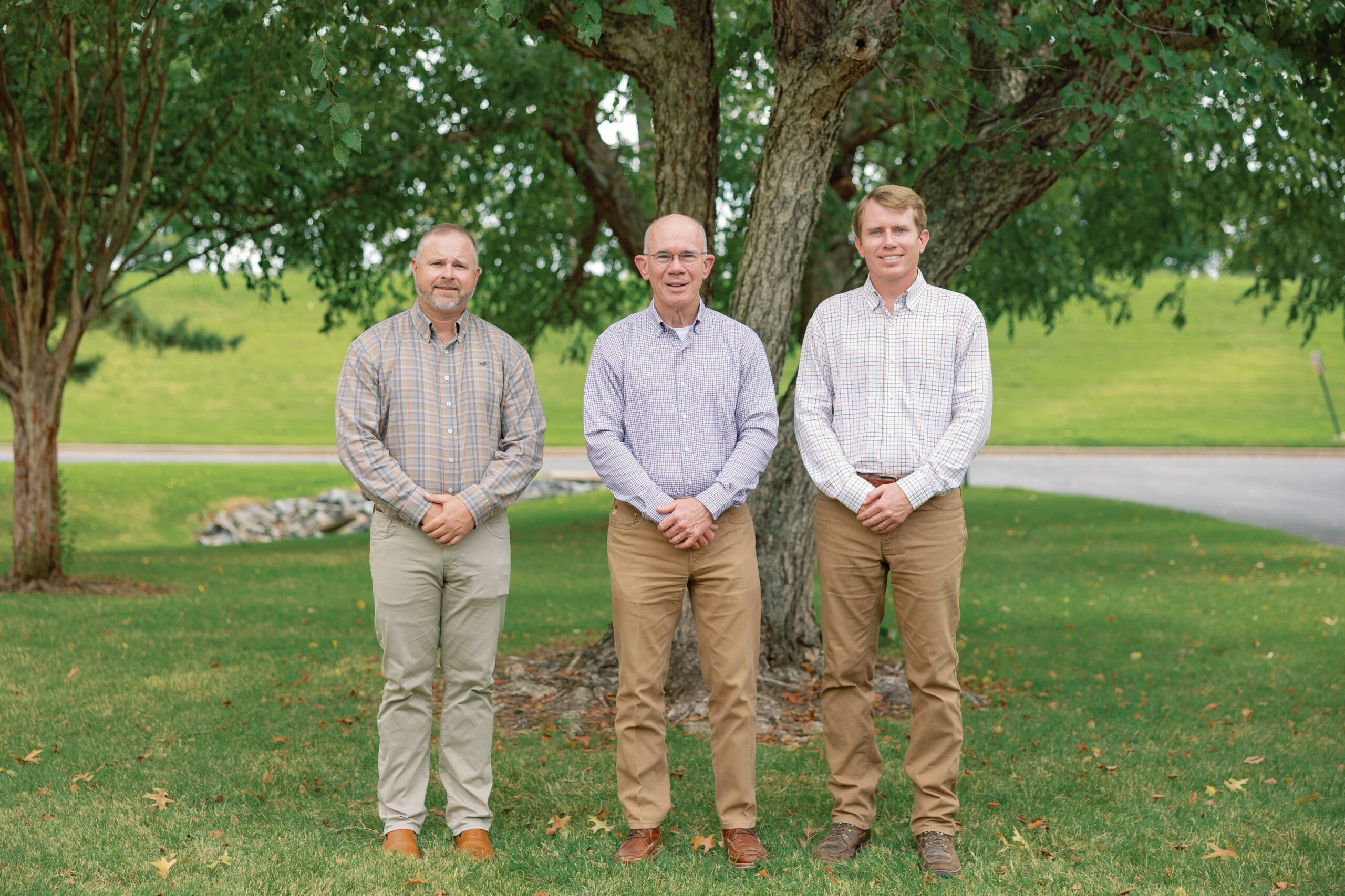 Three men in button-down shirts and khaki pants stand side by side on grass in front of a tree, with a road and green landscape in the background.