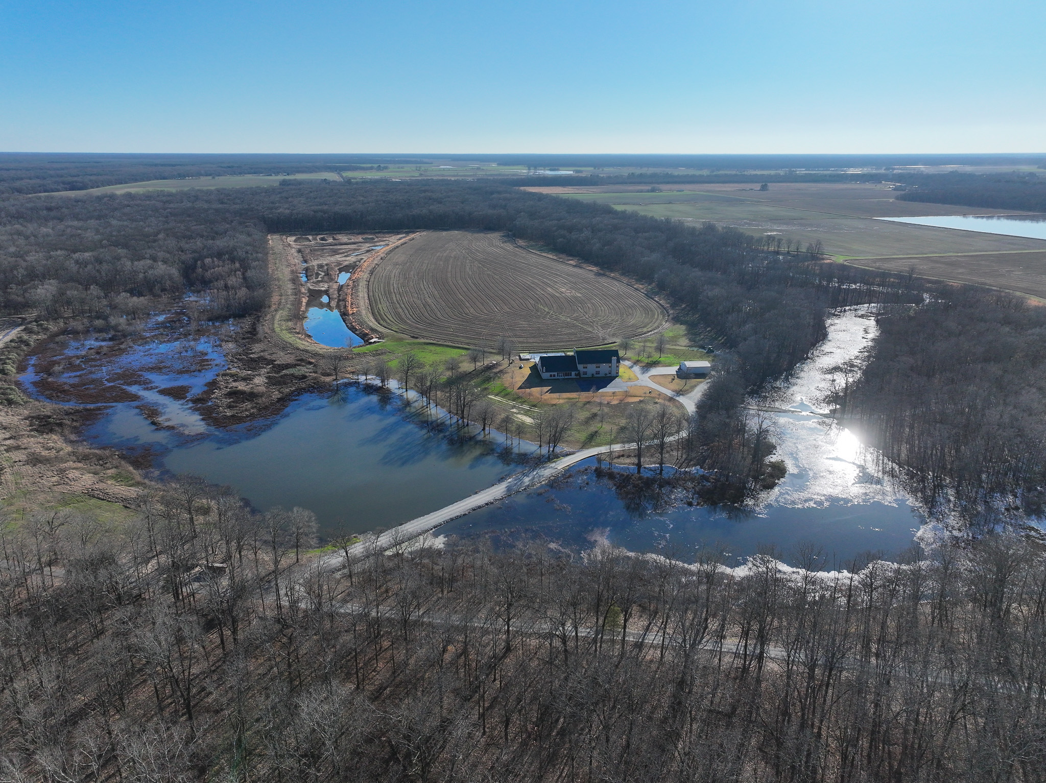Aerial view of a rural farm with a large barn, fields, ponds, and surrounding trees. Sunlight reflects off the water, and a winding road passes through the landscape under a clear blue sky.