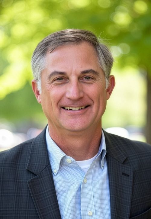 A middle-aged man with short gray hair smiles at the camera. He is wearing a checkered dark blazer over a light blue button-up shirt, standing outdoors with green trees blurred in the background.