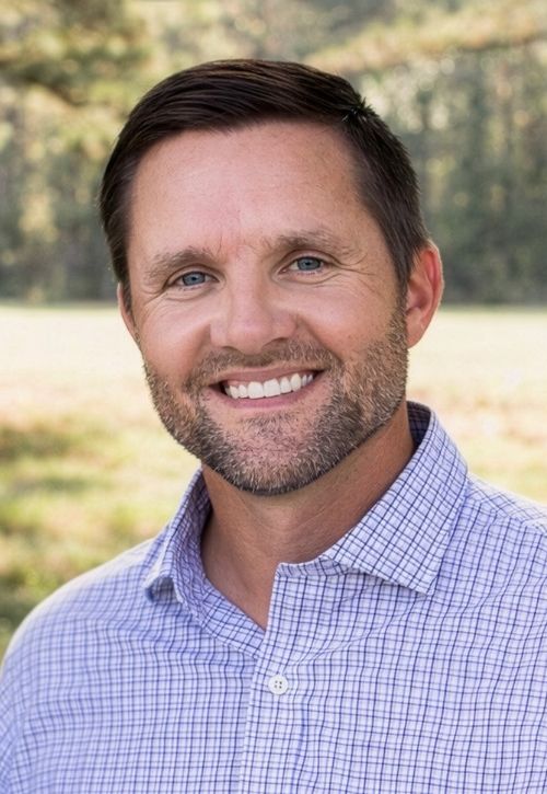A man with short brown hair and a beard, wearing a light purple checkered shirt, smiles outdoors with a blurred background of trees and grass.