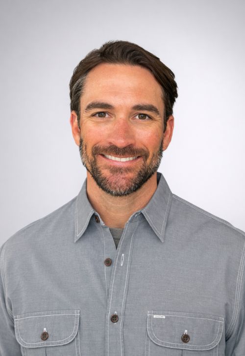 A man with short brown hair, a beard, and a mustache smiles at the camera. He is wearing a light gray button-up shirt and stands against a plain light gray background.