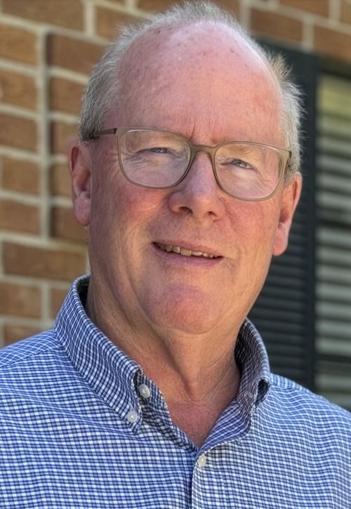 An older man with short gray hair and glasses, wearing a blue and white checkered shirt, stands outdoors in front of a brick wall and a window with shutters, smiling at the camera.