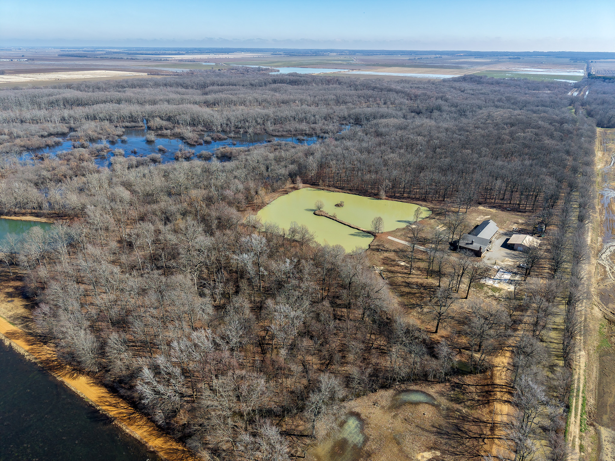 Aerial view of a rural landscape featuring a cluster of leafless trees, several ponds, a farmhouse with outbuildings, and dirt roads, all surrounded by fields and more bodies of water under a clear sky.
