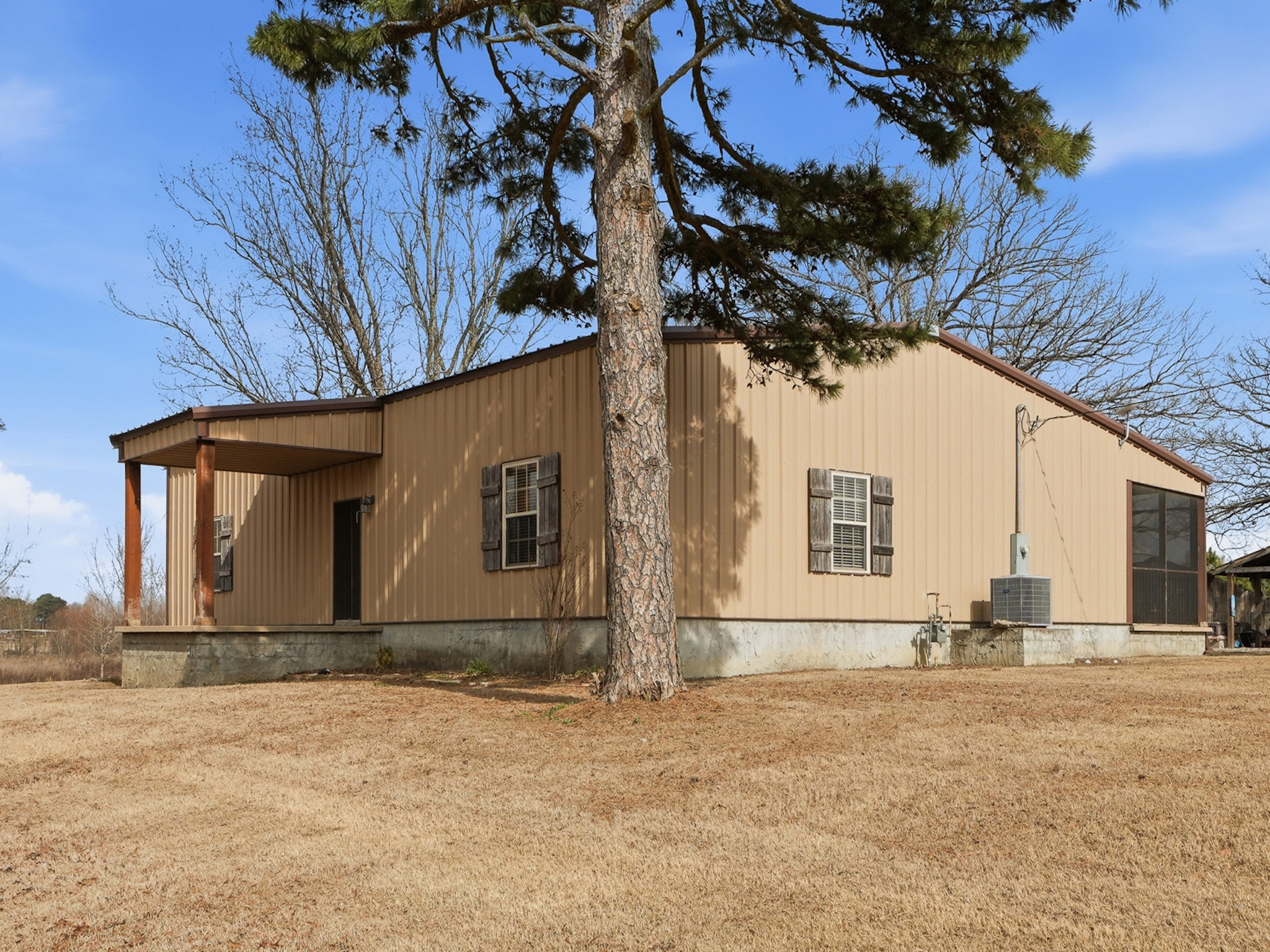 A beige metal-sided house with several windows stands on a grassy, brown lawn. A tall pine tree is in the foreground, and leafless trees are visible behind the house under a clear blue sky.