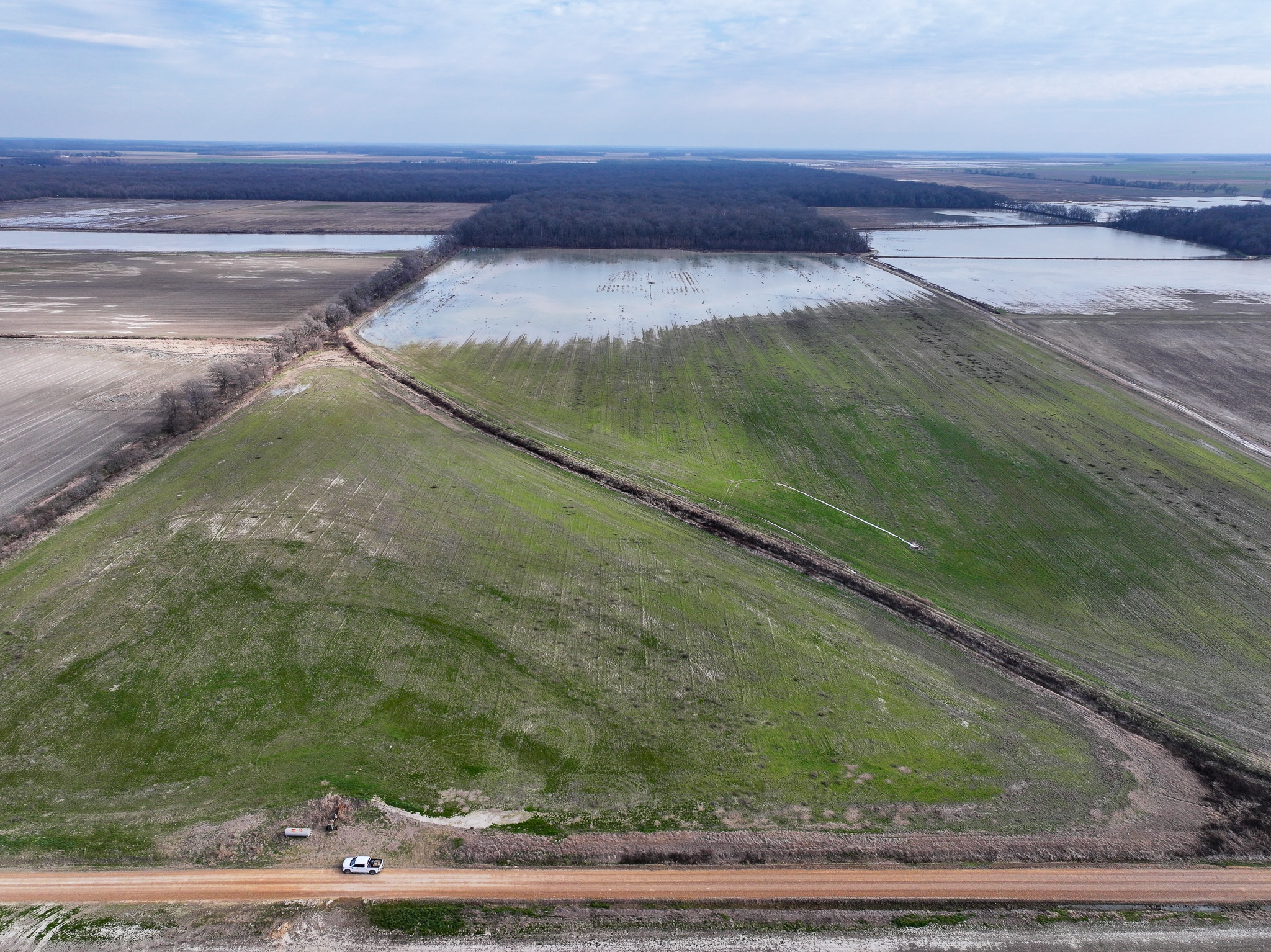 Aerial view of farmland featuring a green triangular field bordered by dirt roads, with adjacent flooded and bare fields. A white vehicle is parked at the field’s edge, and forests are visible in the distance under a cloudy sky.