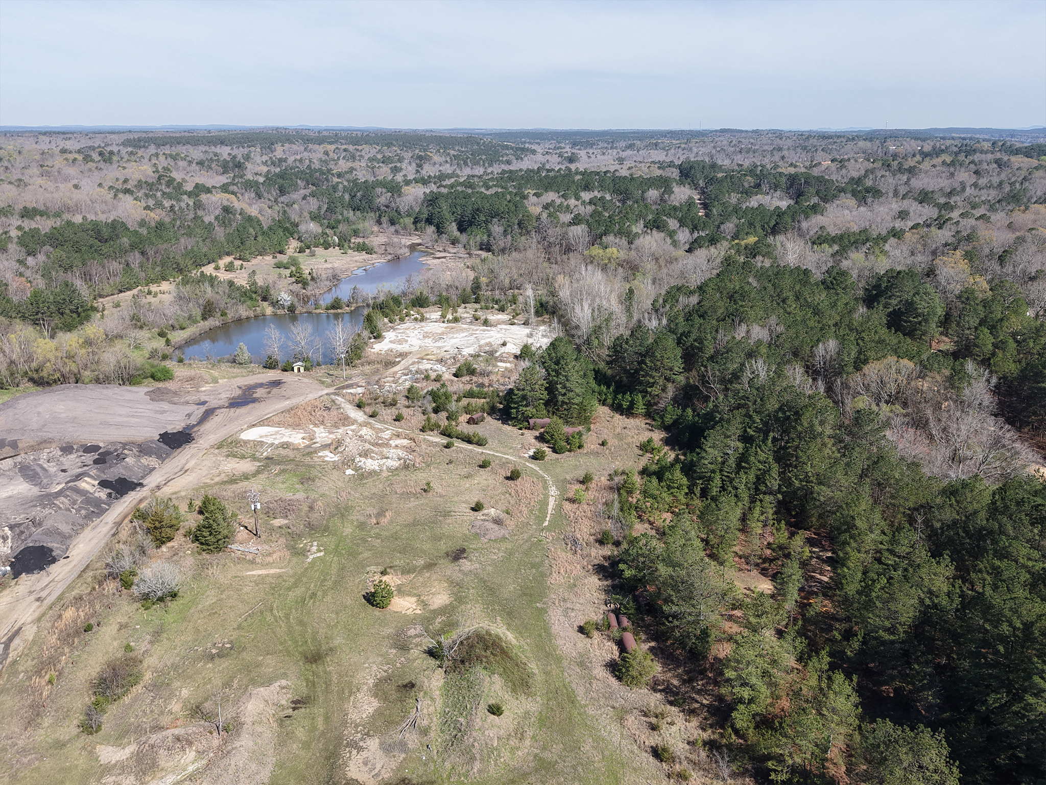 Aerial view of a rural landscape with a pond, patches of forest, open grassy areas, and sandy or rocky clearings under a partly cloudy sky. Dirt paths and scattered trees are visible throughout the scene.
