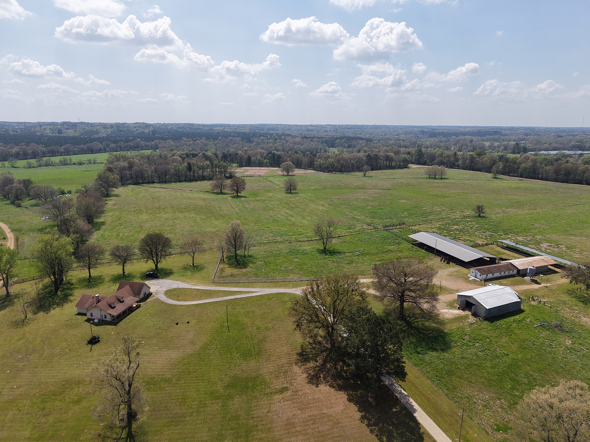 Aerial view of a rural landscape featuring a farmhouse, driveway, several barns, and green fields with scattered trees under a partly cloudy sky.