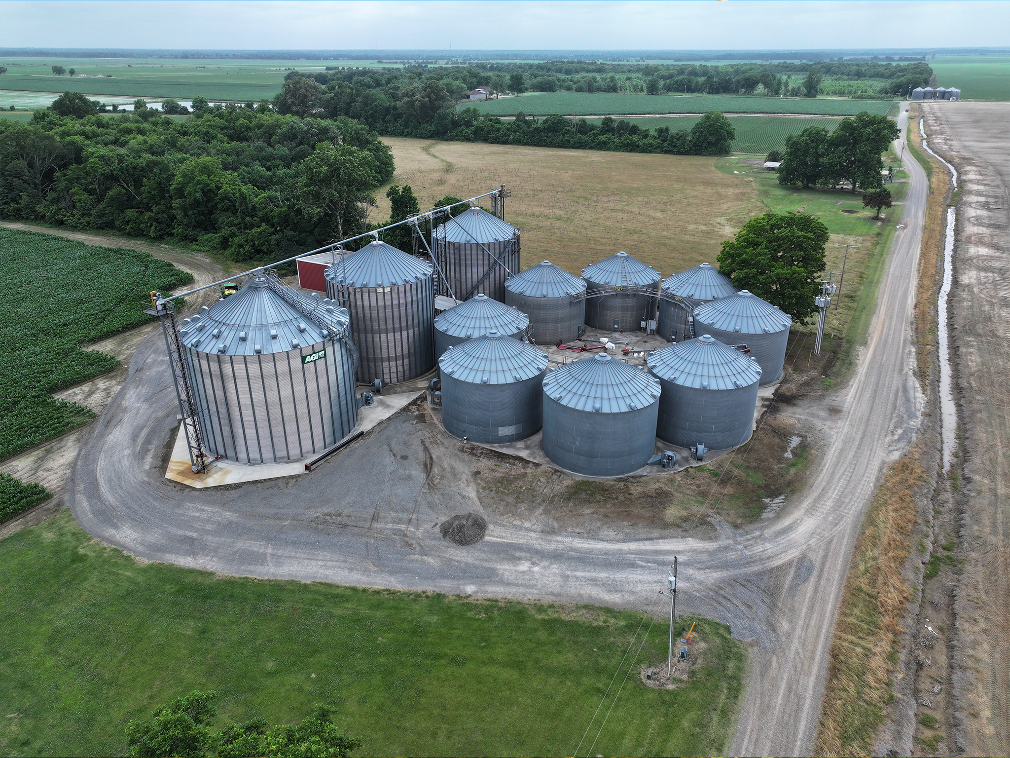 Aerial view of multiple large metal grain silos grouped together on a rural farm, surrounded by green fields, trees, and dirt roads under a cloudy sky.