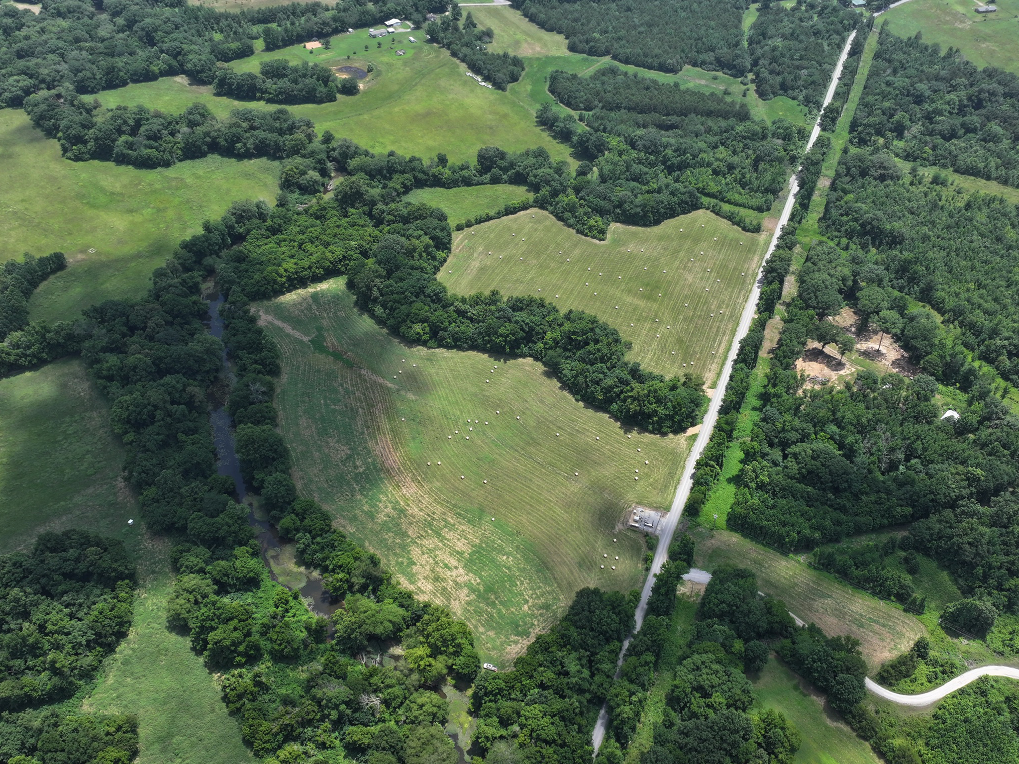 Aerial view of green fields bordered by trees, a winding creek, scattered white dots possibly livestock, and a straight road cutting through the landscape. Some buildings and forested areas are visible.