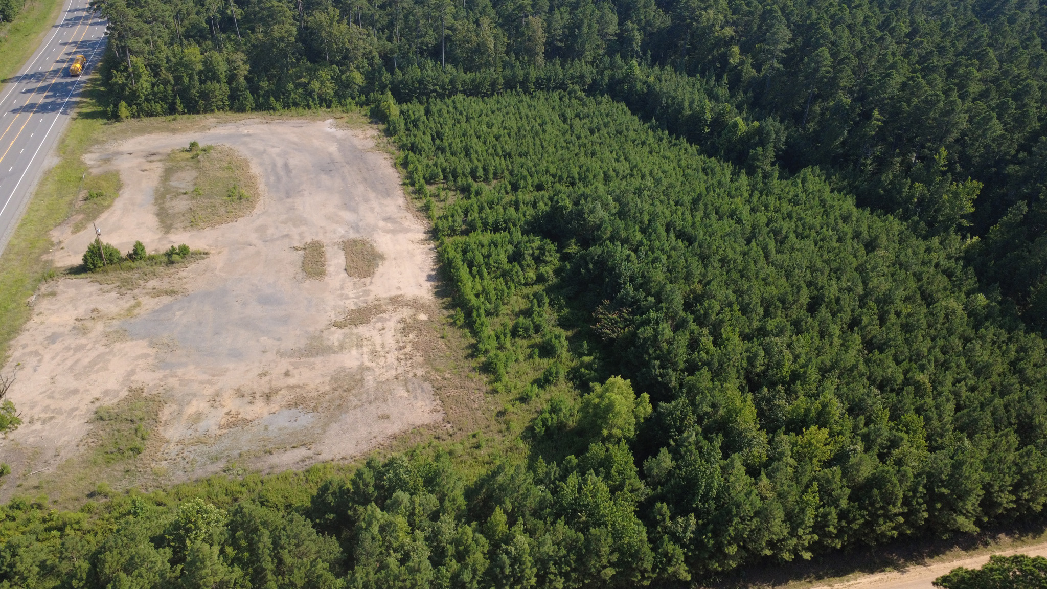 Aerial view of a cleared, empty dirt lot next to a dense green forest, bordered by a road on the left side and surrounded by trees on the other sides.
