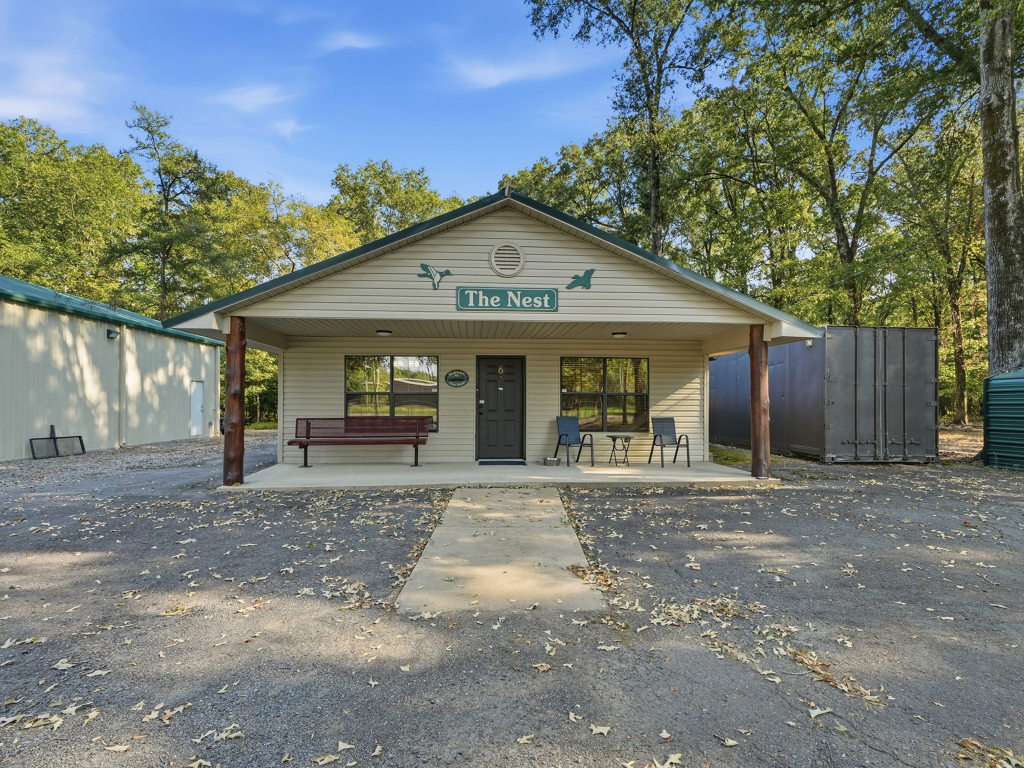 Small building named The Nest with a front porch, bench, and chairs. Surrounded by trees and a paved area with scattered leaves. A storage container and another building are visible nearby.