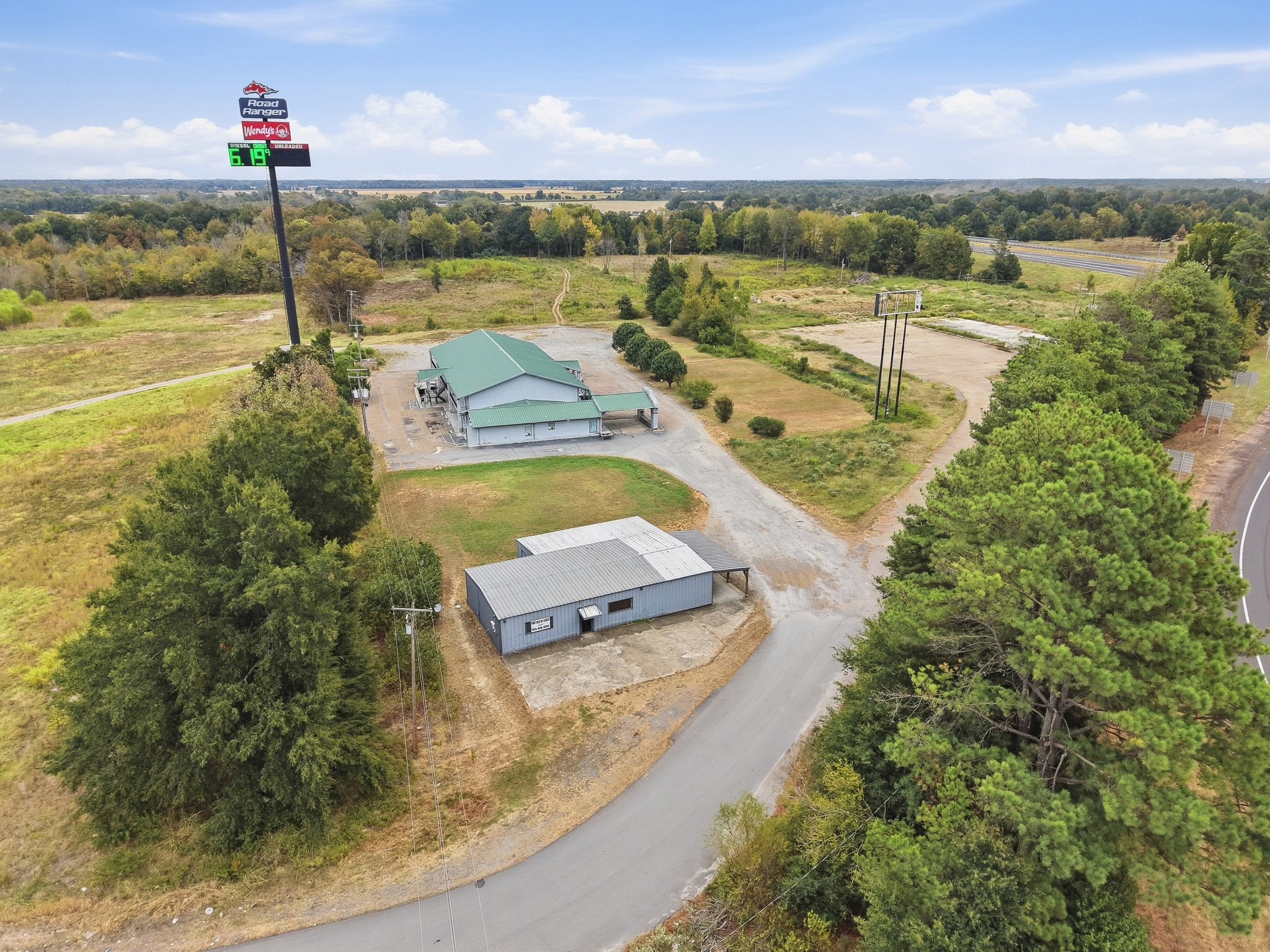 Aerial view of a Road Ranger gas station with a large sign and a separate, smaller metal building located near a highway and forest.